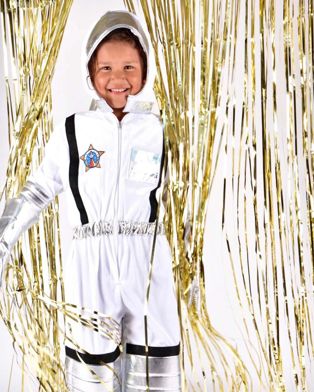 Flicka som bär en vit astronautdräkt med svarta och silvriga detaljer kikar fram genom ett gulddraperi