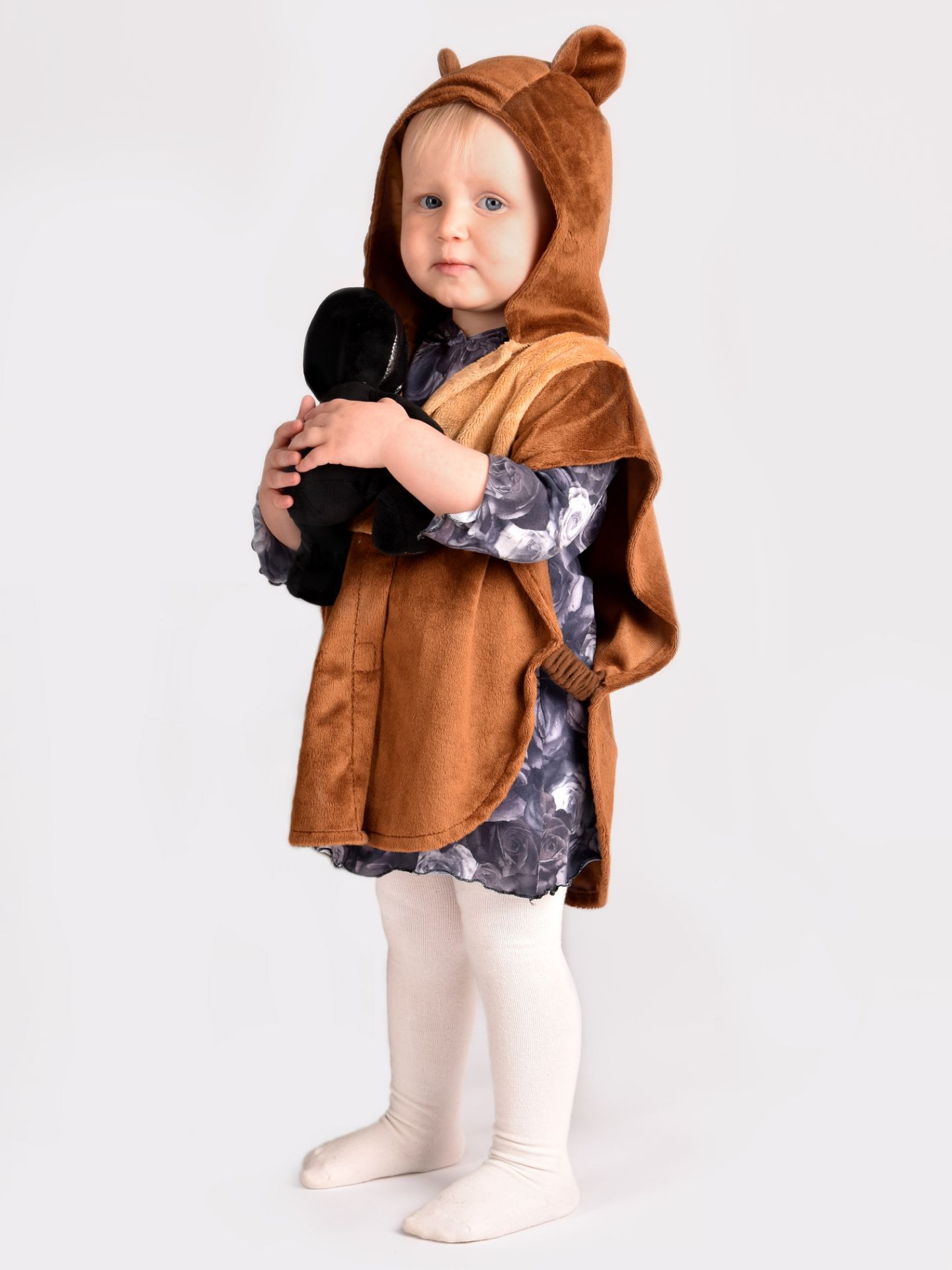 barn som bär en brun björncape med huva med fastsydda öron och rund svans bak. barnet håller i ett svart gosedjur.