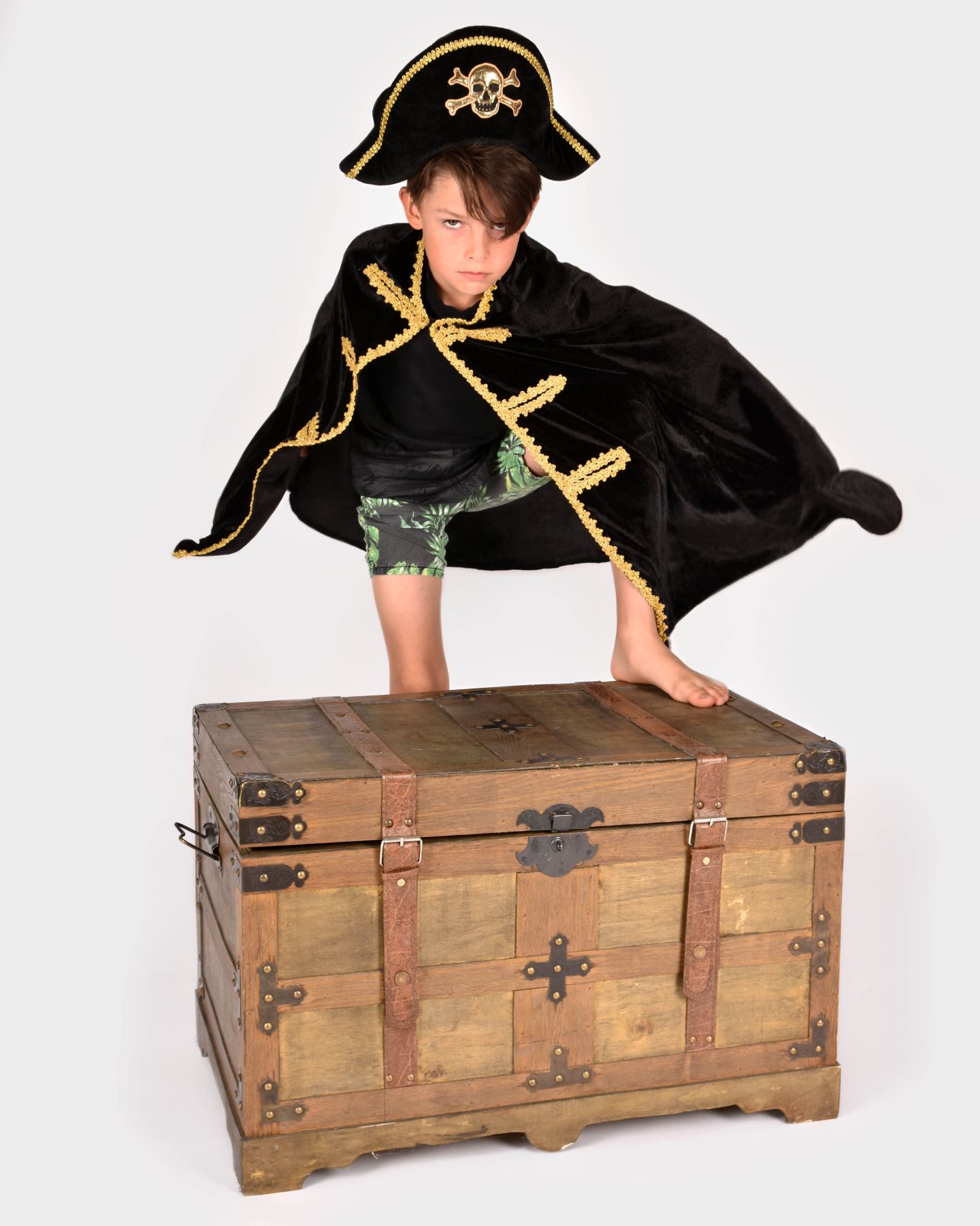 Pojke iklädd en piratcape och pirathatt, båda med gulddetaljer. Pojken står lutad mot en skattkista.
