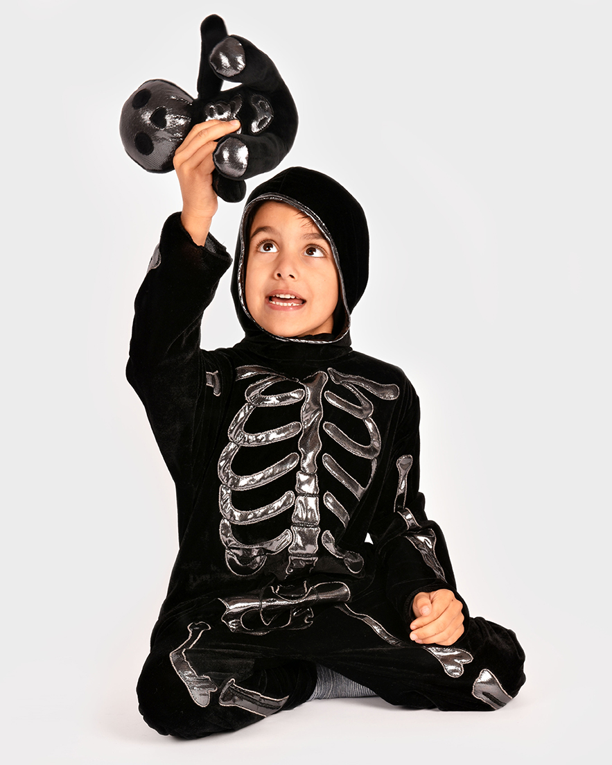 sittande pojke som bär svart skelett-overall med silverdetaljer och håller i ett matchande svart gosedjur med silverdetaljer