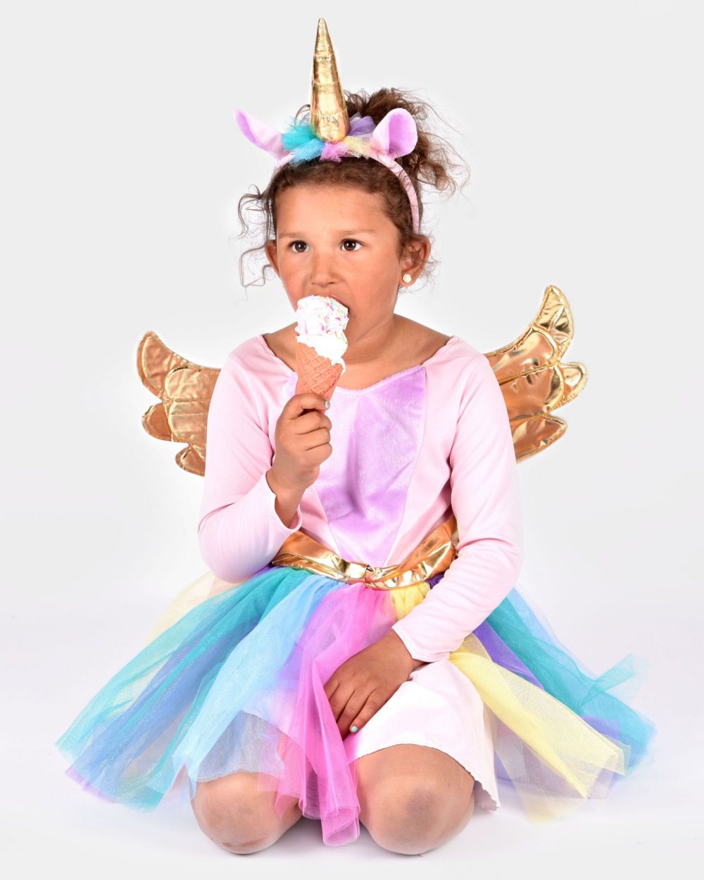Barn som äter glass och bär en multifärgad enhörningsdräkt samt tillhörande diadem med guldigt spiralhorn.