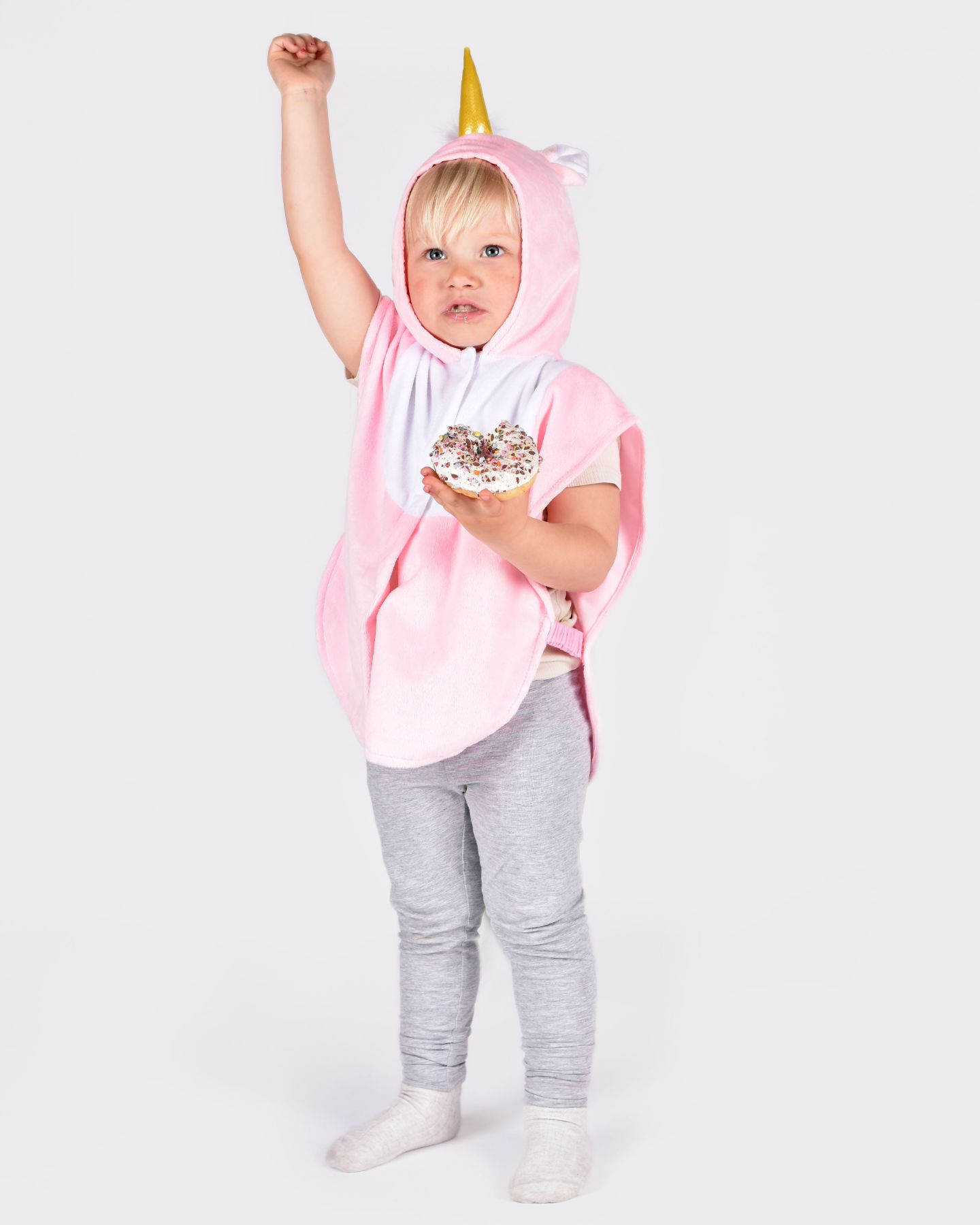 barn som bär en rosavit enhörningscape med öron och ett gyllene horn på huvan. barnet håller en donut i ena handen och den andra handen upp.