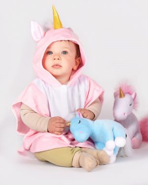 sittande barn som bär en rosavit enhörningscape med öron och ett gyllene horn på huvan. i bild syns också två mindre enhörningsgosedjur i ljusblått och vitt.
